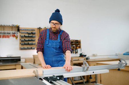 Foto de Carpintero joven que trabaja en sierra circular y se dedica a hacer madera en blanco en el taller - Imagen libre de derechos
