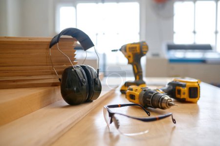 Photo for Carpentry workplace with protective gears and tools for working. Electric drill, protective headset and plastic goggles, wooden timber planks - Royalty Free Image