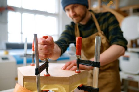 Téléchargez les photos : Jeune menuisier mâle fixant la construction en bois pour la conception intérieure ou l'assemblage de meubles à l'aide de différents outils sur fond d'atelier - en image libre de droit