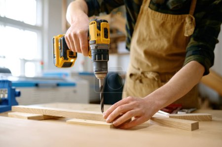Foto de Carpintero trabajando con taladro apoyado sobre mesa en taller de carpintería. Reparador ensamblando piezas de madera usando instrumento profesional - Imagen libre de derechos