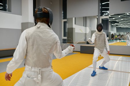 Foto de Dos espadachines con uniforme y casco protector peleando duelo durante el entrenamiento en la sala de gimnasia deportiva. Deportistas en club de esgrima - Imagen libre de derechos