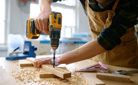 Photo for Carpenter working with drill leaning over table at carpentry workshop. Repairman assembling wooden parts using professional instrument. Closeup view - Royalty Free Image