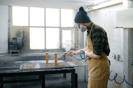 Photo for Young man carpenter wearing protective clothes varnishing board at workshop. Craftsman in respirator mask using professional equipment at home carpentry studio - Royalty Free Image