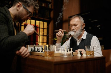 Foto de Familia de gente inteligente jugando al ajedrez, fumando cigarros y bebiendo whisky. Hombres mayores y más jóvenes sentados en la mesa mirando al tablero de ajedrez. - Imagen libre de derechos
