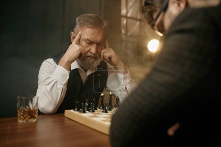 Foto de Hombre mayor ajedrecista tocando templos sintiéndose perdido en pensamientos mientras planea movimiento táctico para luchar contra oponente joven - Imagen libre de derechos