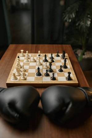 Téléchargez les photos : Table en bois dans la chambre avec des gants de boxer en cuir noir et un échiquier avec des chesspieces. Concept de compétition d'échecs - en image libre de droit