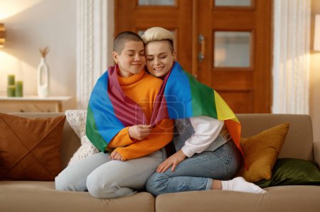 Téléchargez les photos : Jeune femme lesbienne liberté câlins dans lgbt drapeau arc-en-ciel tout en étant assis sur le canapé à la maison salon - en image libre de droit