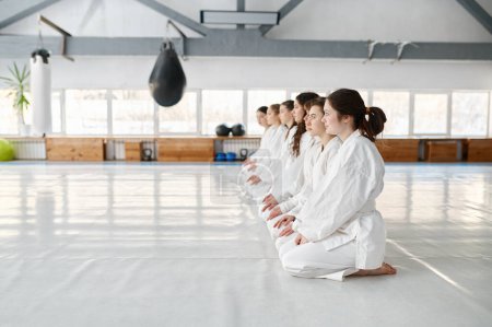 Téléchargez les photos : De jeunes étudiants en aikido portant du kimono assis en rang. Les enfants sportifs adolescents attendent le début de la leçon de groupe ou écoutent leur entraîneur pendant l'entraînement - en image libre de droit