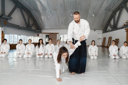 Téléchargez les photos : Instructeur d'Aïkido enseignant les combattants adolescents pendant l'entraînement de groupe dans la salle de gym de l'école de sport. Maître et fille des arts martiaux en kimono montrant la technique de protection pour les étudiants - en image libre de droit