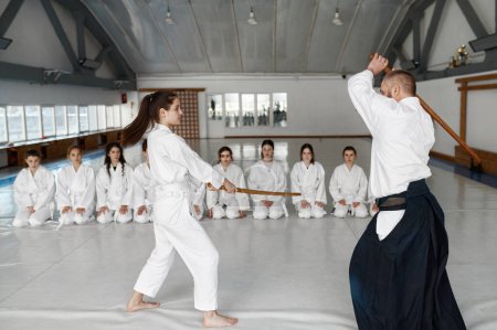 Foto de Maestro de Kendo y pelea repentina con espada de madera bokken durante la clase de artes marciales. Adolescente grupo femenino sentado cerca en el suelo y mirando - Imagen libre de derechos