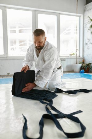 Photo for Mature aikido sensei folding kimono hakama after martial arts training class at gym of professional sport school - Royalty Free Image