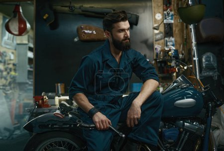 Photo for Portrait of confident man mechanic wearing overalls holding wrench in hand while sitting on repaired motorcycle. Workshop service concept - Royalty Free Image