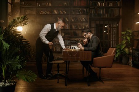 Photo for Two nervous chess players looking at chessboard over cozy home interior. Senior and adult gentlemen having interesting friendly match smoking cigar and drinking scotch - Royalty Free Image
