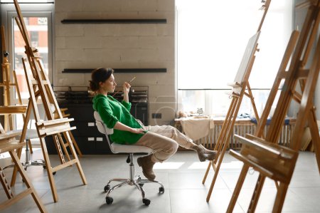 Photo for Female artist with paintbrush in hand looking seriously on her own picture while sitting on chair in art studio - Royalty Free Image
