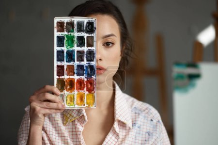 Téléchargez les photos : Portrait de jeune femme artiste couvrant le visage de peintures à l'aquarelle. Peintre créative regardant sérieusement à la caméra. Concept de créativité et d'individualité - en image libre de droit
