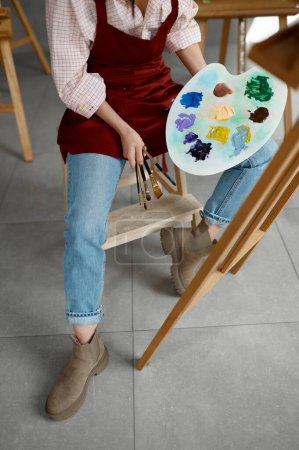 Foto de Foto recortada de artista femenina con delantal sosteniendo pincel y paleta de mezcla de colores sentado frente al caballete de madera. Ambiente de clase de arte hobby creativo - Imagen libre de derechos