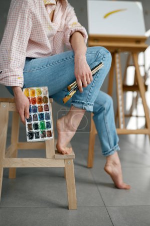 Photo for Cropped shot of barefoot female artist holding drawing tools, watercolor paints palette while sitting on chair at home art workspace - Royalty Free Image