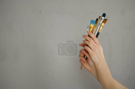 Photo for Female hand holding bunch of paintbrush over grey cement studio wall. Basic supplies for artist and art class school courses advertisement concept - Royalty Free Image