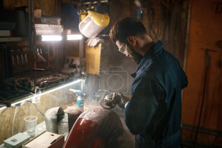 Foto de Reparador puliendo la parte del cuerpo de la motocicleta de la pintura vieja usando la máquina de pelar. Servicio de trabajo en el taller por la noche - Imagen libre de derechos