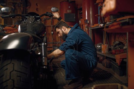 Foto de Mecánico trabajando con motor de motocicleta en taller. Técnico en ropa de trabajo azul sentado vehículo cercano - Imagen libre de derechos