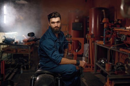 Photo for Portrait of tired but happy workman sitting on repaired motorcycle in workshop. Young adult bearded repairman looking at camera - Royalty Free Image