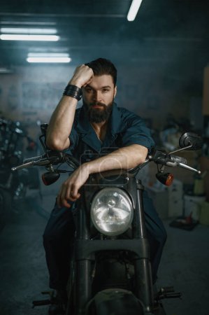 Photo for Portrait of brutal unshaven man mechanic sitting on repaired motorcycle over workshop background - Royalty Free Image