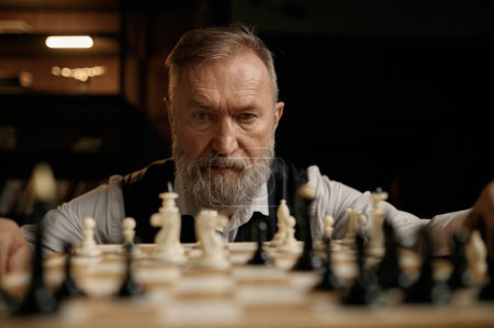 Foto de Retrato facial de un hombre mayor confiado mirando piezas de ajedrez a bordo durante un partido amistoso de competición - Imagen libre de derechos