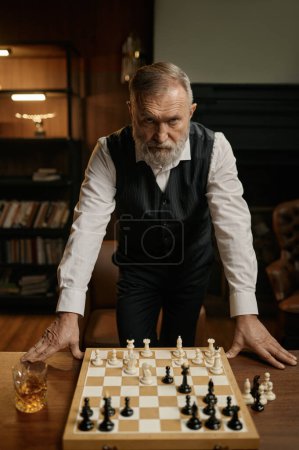 Foto de Retrato de ajedrecista senior serio sobre tablero de ajedrez con pieza de juego. Anciano jugador masculino buscando furiosamente sintiendo riesgo de perder - Imagen libre de derechos
