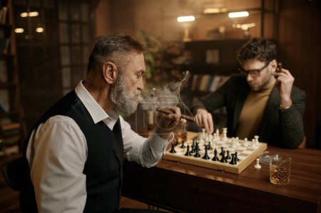 Téléchargez les photos : Deux hommes jouant aux échecs assis à table dans le salon de la maison. Père aîné et fils adulte passent la soirée ensemble à boire du whisky et à fumer des cigares. Jeux intelligents - en image libre de droit