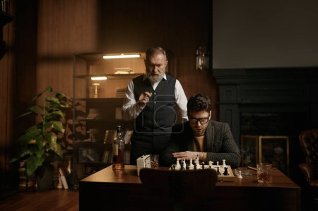 Téléchargez les photos : Deux joueurs d'échecs nerveux regardant l'échiquier sur l'intérieur confortable de la maison. Messieurs âgés et adultes ayant match amical intéressant fumeurs cigares - en image libre de droit
