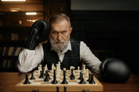 Photo for Serious senior man chess player sitting at chessboard wearing boxer gloves. Portrait shot - Royalty Free Image
