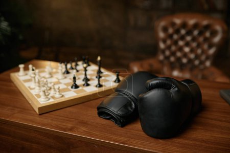 Téléchargez les photos : Table en bois dans la chambre avec des gants de boxer en cuir noir et un échiquier avec des chesspieces. Concept de conflit d'échecs - en image libre de droit
