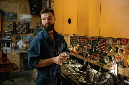 Photo for Portrait of young bearded serviceman looking aside standing over garage workshop interior. Tired mechanic wipes hands with rag after repair work on motorcycle - Royalty Free Image