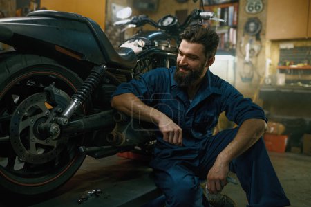 Photo for Satisfied smiling professional mechanic feeling glad looking at repaired motorcycle after good work in repair workshop - Royalty Free Image