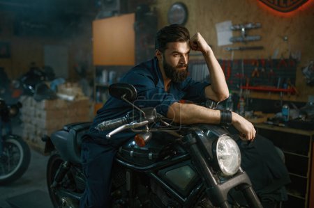 Photo for Portrait of brutal unshaven man mechanic sitting on repaired motorcycle over workshop background - Royalty Free Image