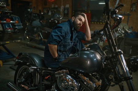 Foto de Retrato del hombre mecánico brutal sentado en la motocicleta reparada sobre el fondo del taller - Imagen libre de derechos