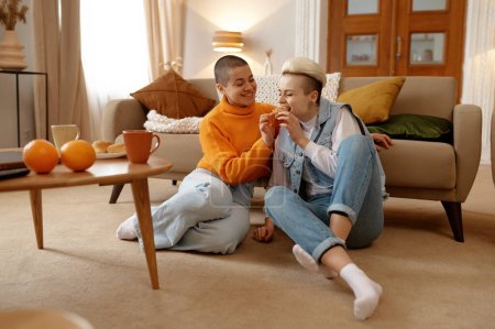 Photo for Happy lgbt woman couple eating taste snacks while sitting at home living room. Cheerful lesbian girlfriends enjoying time tougher on weekend - Royalty Free Image