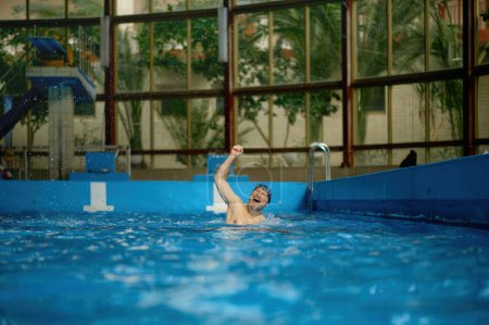 Téléchargez les photos : Homme âgé excité levant joyeusement la main vers le haut se réjouissant de la victoire dans les compétitions de course de natation pour les personnes âgées à la piscine intérieure - en image libre de droit