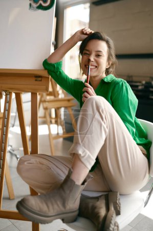 Téléchargez les photos : Portrait d'une jeune artiste souriante ravie tenant un pinceau sur le nez posant pour une caméra à proximité de la toile - en image libre de droit