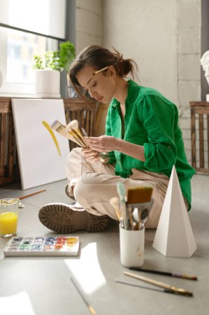 Photo for Pretty young female artist drawing picture on canvas with oil paints sitting on floor in her own art studio. Creativity in work and contemporary creative painter concept - Royalty Free Image