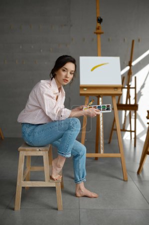 Photo for Young woman artist with creative accessories sitting on chair at artwork workplace with wooden easel on background - Royalty Free Image