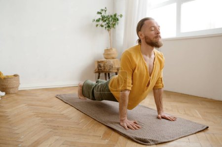 Foto de Joven atlético de pie en la pose de yoga Urdhva Mukha Svanasana. Hombre deportista practicando perro boca arriba. Capacitación en el hogar para la salud y el cuidado corporal - Imagen libre de derechos