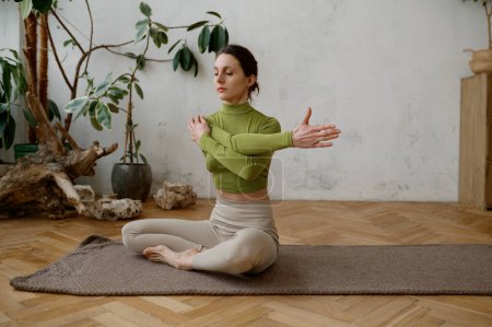Foto de Mujer activa joven haciendo ejercicio mientras está sentada en la esterilla de yoga estirando los brazos en la sala de estar en casa - Imagen libre de derechos