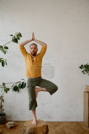Téléchargez les photos : Homme yogi fort équilibrage sur souche en bois dans la pose de yoga arbre Vrikshasana tenant la main dans un geste namaste sur la tête - en image libre de droit