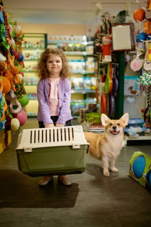 Photo for Portrait of little girl with her corgi dog at pet shop. Little child standing with new transportation carrier for puppy - Royalty Free Image