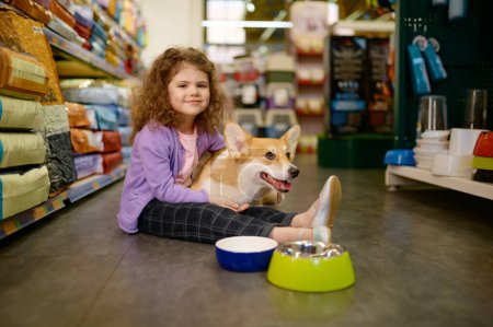 Photo for Little girl child sitting on pet shop floor holding corgi dog on knees. Shopping at supermarket offering goods for domestic animals - Royalty Free Image