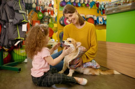 Young mother and little daughter choosing blanket for their corgi dog at pet shop. Happy family spending time together at supermarket for domestic animal