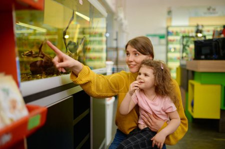 Photo for Excited mother and daughter looking on swimming fish in aquarium at pet shop. Happy family choosing new domestic animal at zoo store - Royalty Free Image