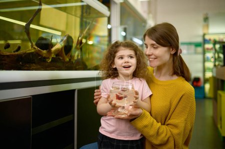 Téléchargez les photos : Portrait de mère heureuse et petite fille a acheté du poisson à animalerie. Focus sur le visage d'enfant émerveillé - en image libre de droit
