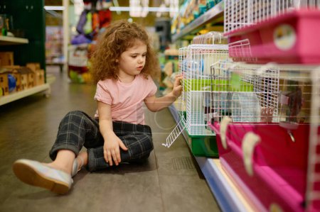 Little girl sitting on pet shop floor nearby cage with rodent. Child dreaming about new pet wishing to buy mouse or hamster